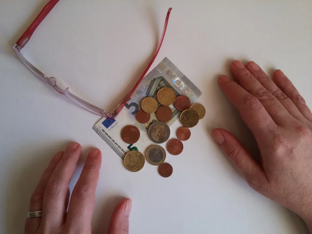 Manos de una persona mayor junto a unas gafas, un billete de cinco euros y monedas